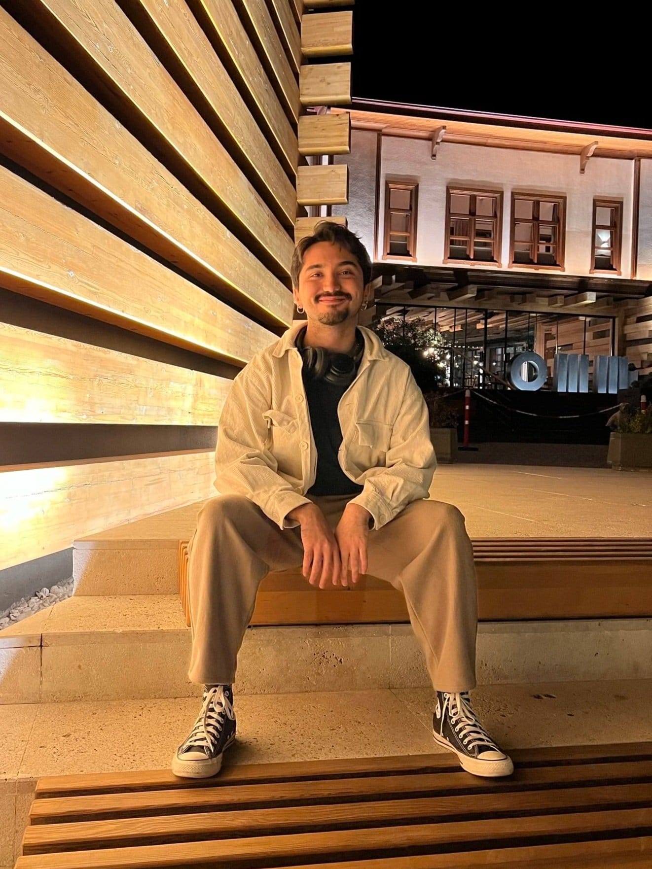 Murat sitting on a wooden bench outside at night, smiling, with lit-up buildings behind him.